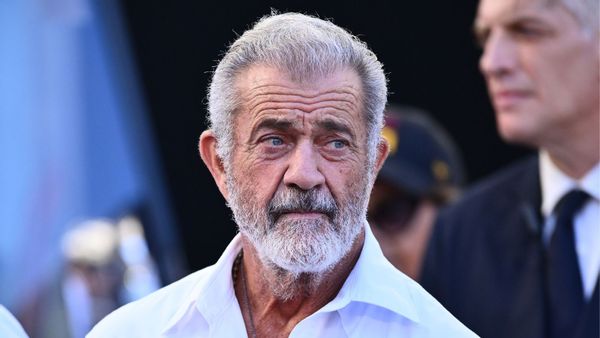 An image shows a photo of actor Mel Gibson with glasses tucked into a partially unbuttoned white collared shirt, with Gibson looking to his left and a man out of focus in the background wearing a suit and tie.