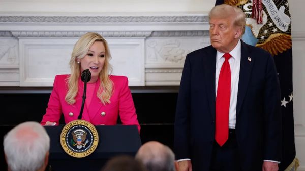Paula White-Cain, a middle-aged white woman with platinum blonde hair, speaks at a podium wearing a neon pink suit. Donald Trump stands to her left, watching.