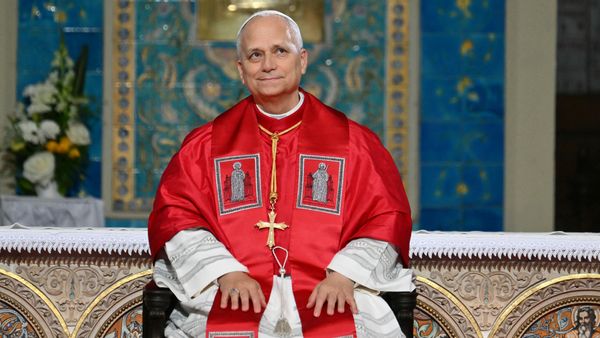 Pope Leo XIV, an older white man wearing a white skullcap and red papal robes, sits on a bench. 