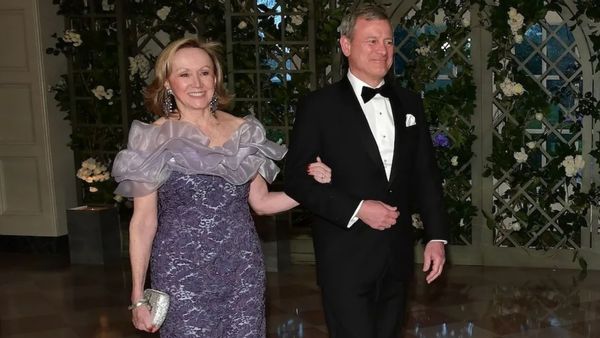 An older white woman with shoulder length blonde hair wearing a purple dress and an older white man wearing a black tuxedo. 