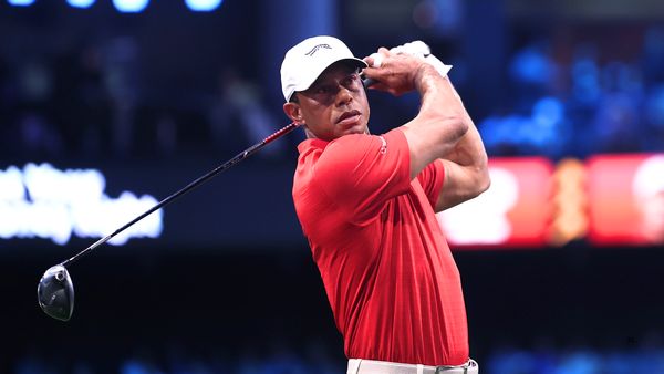 Tiger Woods, a black man wearing a golf cap and a red polo shirt, swings a driver while playing in an indoor golf arena.