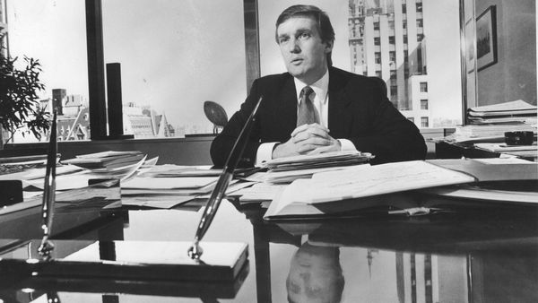 A black and white image of then-New York City landlord Donald Trump, a younger white man with slicked back hair, sitting in a high-rise office. 