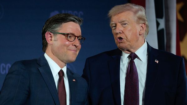 Rep. Mike Johnson, a white man with short graying hair wearing tortoiseshell glasses and a suit, leans in to listen to U.S. President Donald Trump, a white man with white-blond hair also wearing a suit.