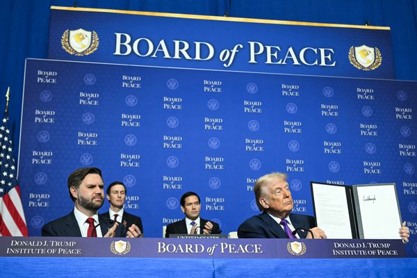 Two white men (U.S. Vice President JD Vance and President Donald Trump) wearing suits, in front of a large blue backdrop reading "Board of Peace." Trump holds up an executive order with his signature on it.