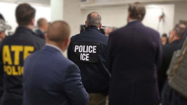 A federal agent stands facing away from the camera wearing a jacket that reads "ICE."