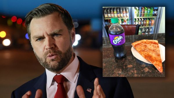 An image shows a fairly close picture of US Vice President JD Vance standing outside in a suit, red tie and American flag pin, with a blurry backdrop of an airport, gesturing with his hands while speaking to journalists.