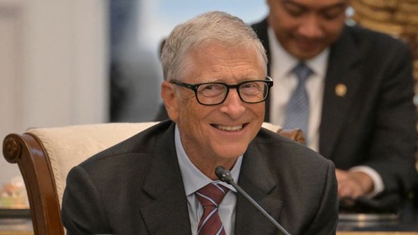 Bill Gates, a white man with dark-framed glasses, smiles as he sits before a microphone.