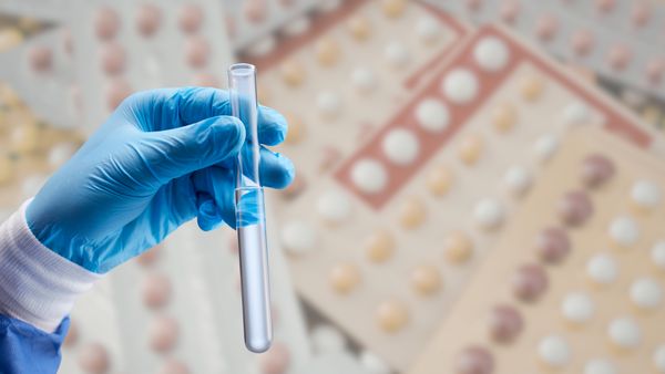 A blue gloved hand holding a test tube in front of a background of birth control pill packs. 
