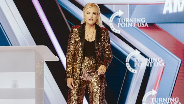 Erika Kirk, a blonde woman in a sequined gold suit with blue eyes, stands on a stage with a pattern of "TURNING POINT USA" logos behind her.