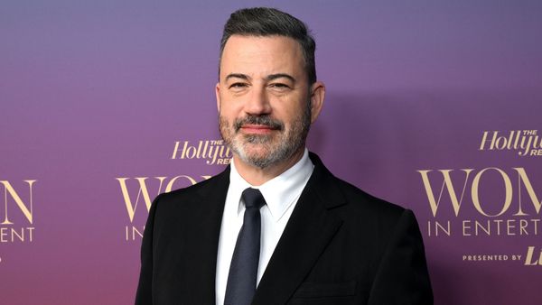 Jimmy Kimmel, a middle-aged white man with short, black hair and a salt-and-pepper beard, wears a black suit in front of a purple background.