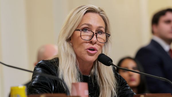 Marjorie Taylor Greene, a middle-aged white woman with long, platinum blonde hair and glasses, wears a black leather jacket while speaking into a microphone.