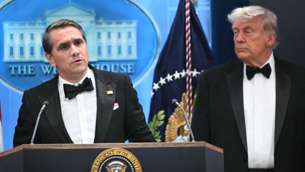 U.S. acting Attorney General Todd Blanche, a middle-aged white man in a black and white tuxedo, speaks behind a podium while U.S. President Donald Trump looks on.