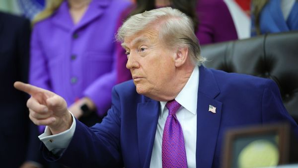 An image shows U.S. President Donald Trump sitting in the Oval Office in the White House pointing while wearing a suit and tie, and American flag pin.