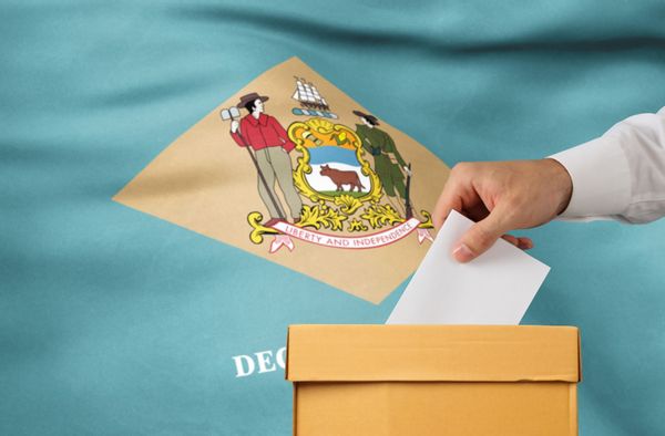 A hand puts a piece of paper in a ballot box in front of the Delaware flag.