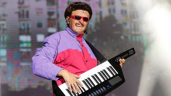 Oliver Tree, a young white man, wears a black, bowl-cut wig, goofy red sunglasses and a neon pink and purple puffer jacket while playing a keytar.