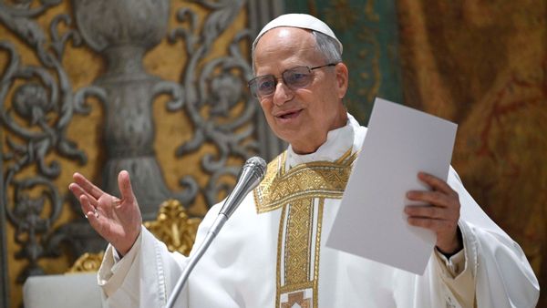 A photo shows Pope Leo XIV wearing a white and gold outfit, glasses and a zucchetto, including while speaking into a microphone and holding paper in his left hand.
