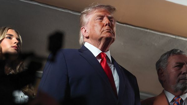 Trump, a white man, stands and stares ahead of him wearing a dark blue suit, a white shirt and a red tie. The face of a young woman, his granddaughter Kai Trump, appears behind him. To his left is Knicks owner James Dolan, a white man with white hair and a salmon jacket.