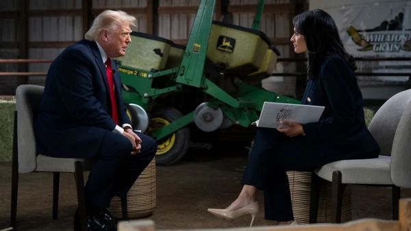 Trump leans forward in his seat while speaking to Kristen Welker. They appear to be sitting in a barn of sorts and a large piece of John Deere farm machinery is visible behind them. 