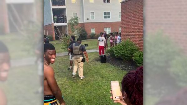 An image shows an early evening setting outdoors at an apartment complex with Black students throwing water balloons, shooting water guns and dumping water on a man appearing to wear a uniform and vest.