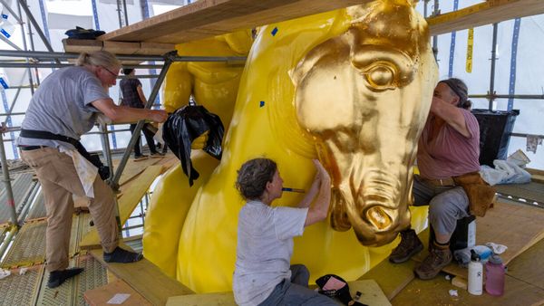 Several women apply gold leaf to a large yellow horse statue that is partially covered in gold leaf. 
