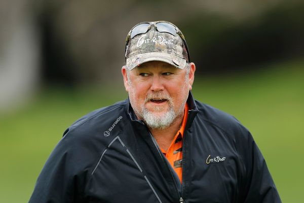Larry the Cable Guy, an old white man with a bristled, white goatee, wearing a camouflage baseball cap with wrap-around sunglasses, and a black windbreaker. 