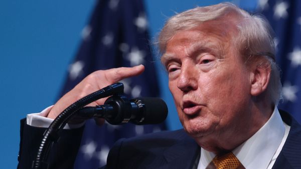 U.S. President Donald Trump raises a finger to his head while speaking in front of a microphone. 