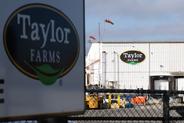 A sign and warehouse displaying the logo of Taylor Farms, a gold-ringed oval with a black interior containing the company's name above a green leaf.