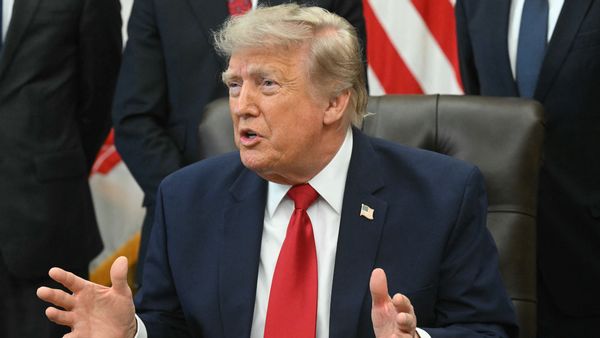 President Trump, a white man with light hair, wears a blue suit jacket, white shirt and red tie. He sits in a black chair while gesturing with both hands.