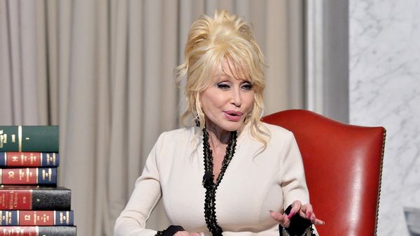 Dolly Parton, a white woman with bright blonde hair, speaking while seated in front of a pile of books.