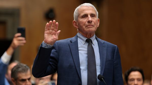 Anthony Fauci, an older white man wearing a blue suit, stands and holds his right hand up to take an oath. 