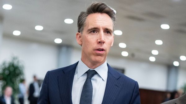 A photo shows a head-to-waist shot of U.S. Sen. Josh Hawley, R-Mo., standing in a well-lit room wearing a blue striped suit, light blue shirt and dark blue tie, with people and a plant visible in the out-of-focus background.