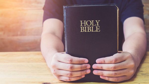 A bright photo shows a person seated in a short sleeve shirt on a wooden bench holding a Bible up on a wooden table with the words "Holy Bible" facing the camera, with the camera pointed to focus on the Bible, each of the person's hands holding part of the bottom of the book, and the person's face and head out of frame and not visible above the image.