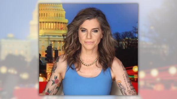 Nancy Mace, a white woman with brown hair, sits in front of a podcast mic in a blue, sleeveless top that shows various tattoos on both her arms.
