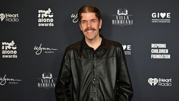 Perez Hilton, a younger middle-aged man with combed, middle parted hair and a mustache, wearing a black leather jacket.