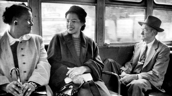 A black-and-white image showing Rosa Parks, a Black woman wearing glasses and a overcoat, sitting on a bus seat beside another Black woman and a white man. 