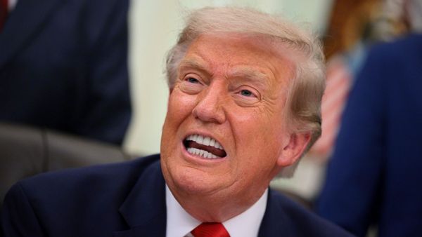A photo shows a close-up view of US President Donald Trump speaking in a dark blue suit, white shirt and red tie while seated in the Oval Office.