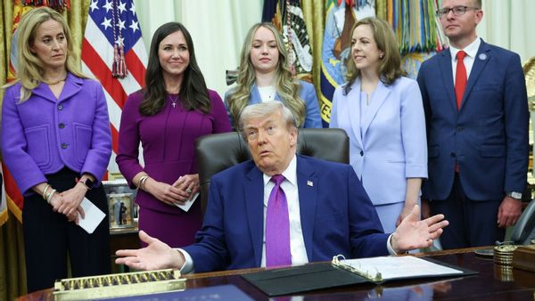 U.S. President Donald Trump, dressed in a blue suit, white shirt and purple tie, sits in a chair behind the Oval Office’s Resolute Desk and gestures with each hand out to each side. Five people stand behind him, including Republican U.S. Senator Katie Britt of Alabama, who wears a purple outfit and a cross necklace.