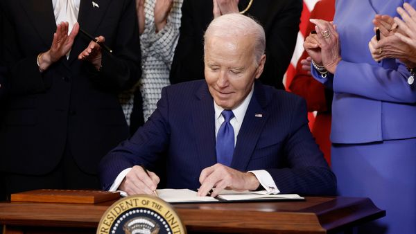 Joe Biden, an older white man wearing a blue suit, sits at a desk and sign a piece of paper as a crowd of people stand behind him looking on.