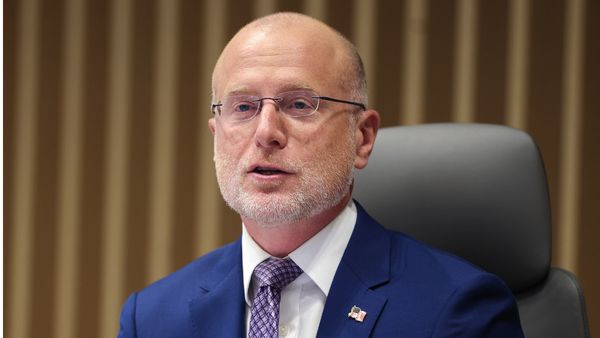 Brendan Carr, an older but still middle-aged White man, sits in a chair. He is bald, and wears a blue suit and glasses.