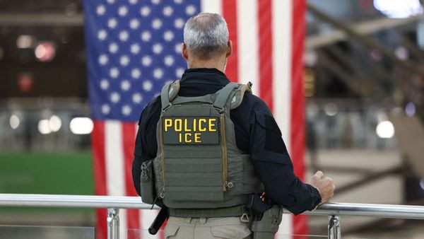 A photo shows a man with short gray hair and some balding wearing a U.S. Immigration and Customs Enforcement vest displaying the words "POLICE" and "ICE," as well as carrying equipment including a firearm, facing away from the camera and looking down from a railing toward an airport-like scene, complete with a very large draped U.S. flag in the background.