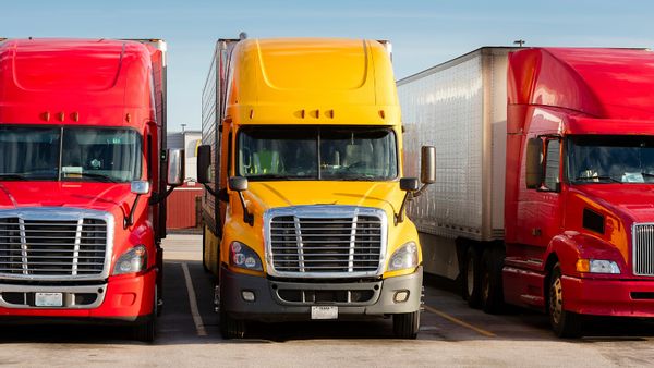 An image shows the front of three red or yellow semi-trucks parked next to each other in a sunny parking lot, with a building barely visible in the background displaying a McDonald's arches sign.