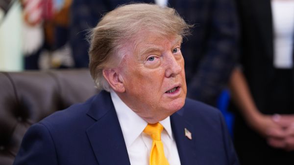 A man in blue suit with yellow tie and American flag pin sitting in a chair and looking off camera with his mouth open slightly.