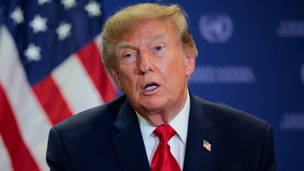 U.S. President Donald Trump wears a suit and red tie and sits in front of a blue background featuring an American flag.