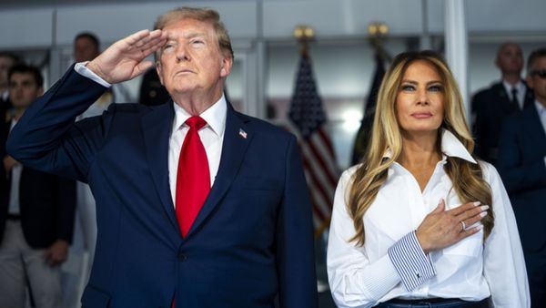 U.S. President Donald Trump salutes while standing next to first lady Melania Trump, who holds a hand up to her heart. 