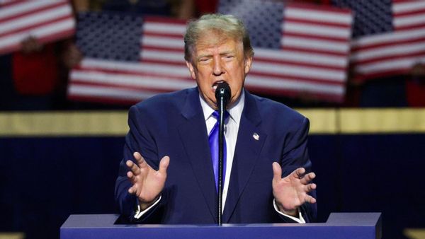 A photo shows U.S. President Donald Trump wearing a blue suit, blue tie and white collared shirt with an American flag pin, gesturing with both hands while standing behind a blue lectern, in front of out-of-focus people holding American flag signs in the background.