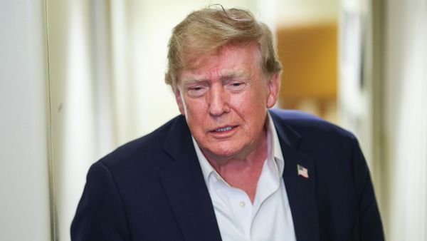A photo shows U.S. President Donald Trump wearing a blue blazer, white shirt with top two buttons unbuttoned, a U.S. flag pin and a very slightly smiling facial expression as if he's listening to a journalist ask a question while leaning forward, appearing on Air Force One.