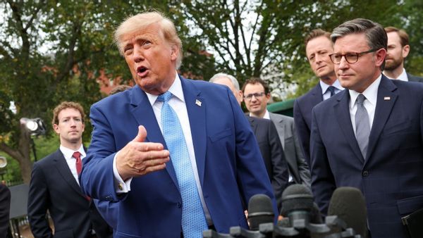 This photo shows an outdoor scene at the White House with U.S. President Donald Trump wearing a blue suit, blue tie, white shirt and U.S. flag pin, while gesturing and speaking in front of microphones with tech leaders and House Speaker Mike Johnson, who is wearing a dark suit, gray tie, white shirt and lapel pin, as well as background appearances by Meta CEO Mark Zuckerberg and others.