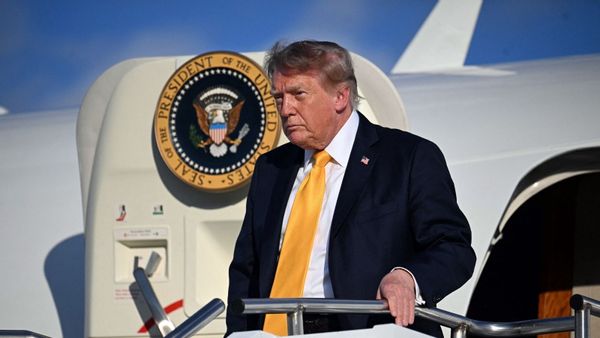 An image shows U.S. President Donald Trump wearing a dark suit, white shirt, yellow tie and U.S. flag pin while standing on the stairs outside the door to the plane known when he's onboard as Air Force One.