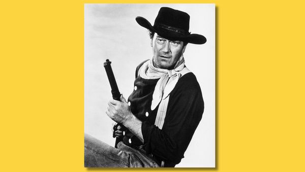 Black-and-white image shows actor John Wayne seated in a cowboy hat while holding up a gun. 