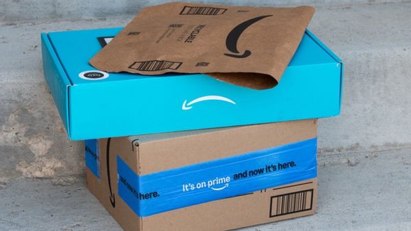 A stack of brown and blue cardboard boxes and packages with Amazon logos sit on a porch. 